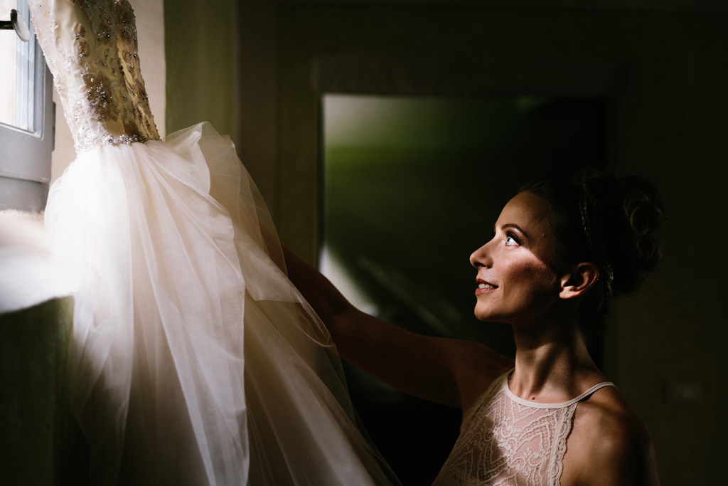 Fotografo Matrimonio Convento Dell'Annunciata Medole La sposa prende il suo abito bianco appeso alla finestra illuminata da una luce splendida